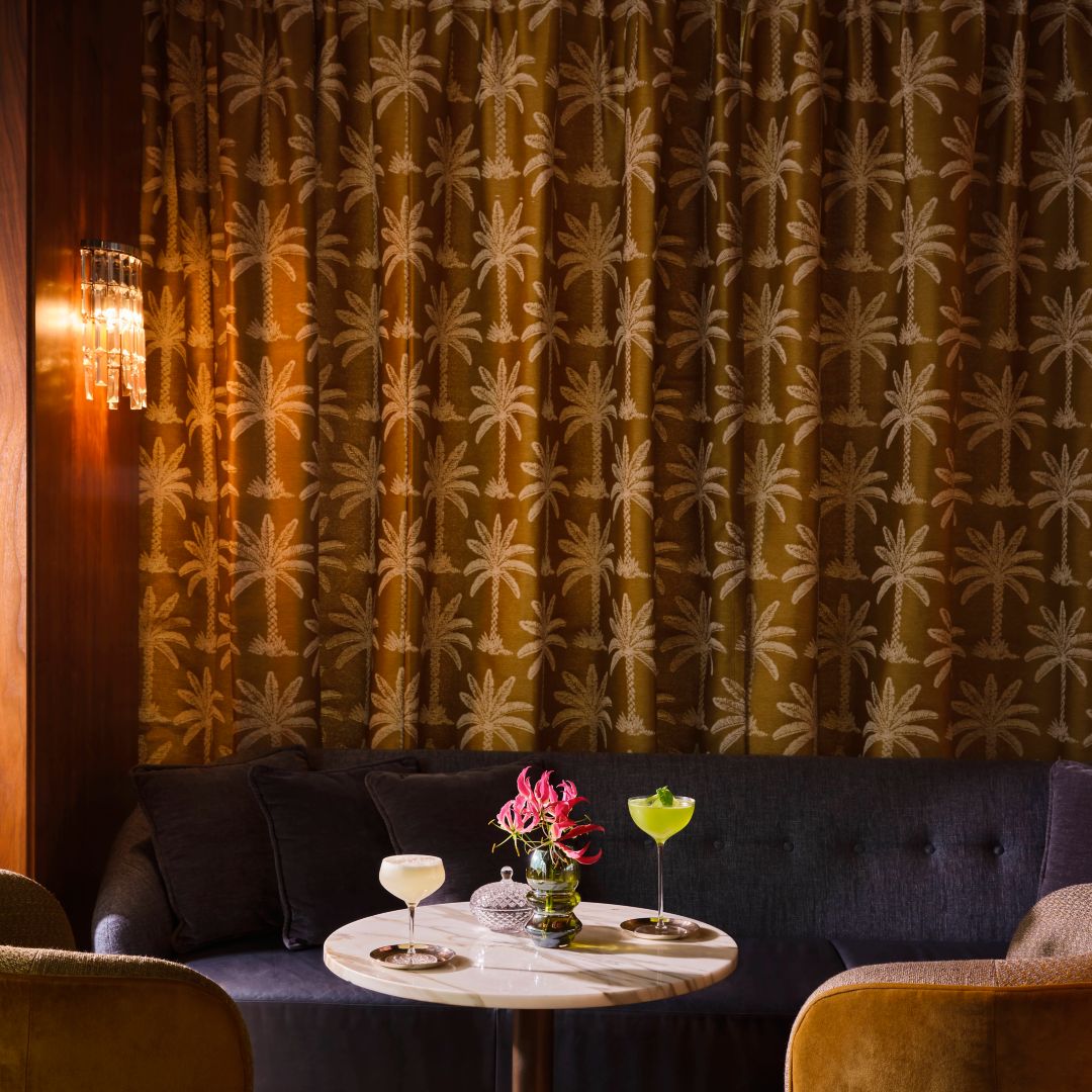 A speakeasy ambiance at THE1835: Two drinks and Gloriosa lilies adorn the round marble table, while patterned curtains lend the room a warmly inviting atmosphere.